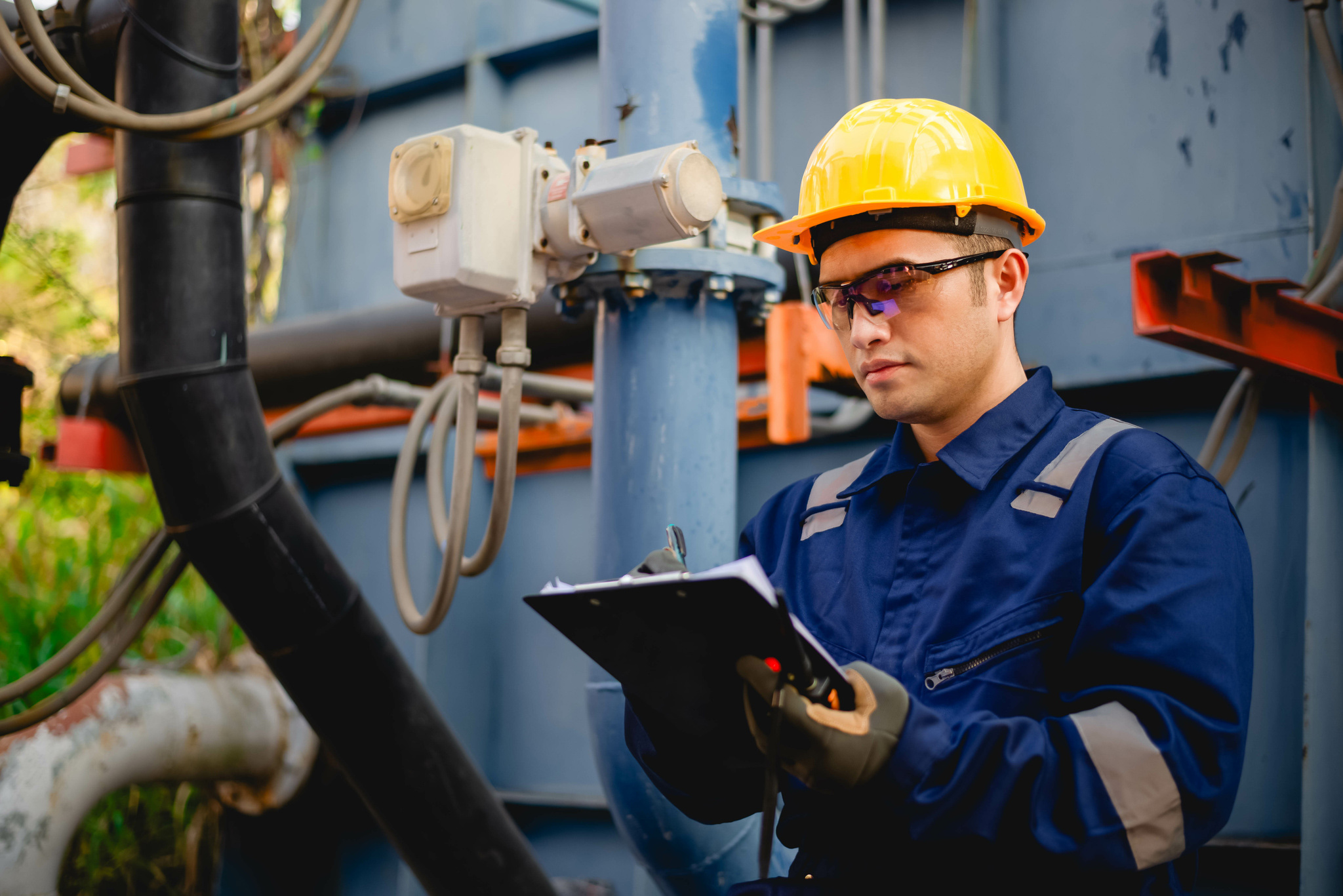 A man in a hardhat is taking notes as he looks at pipes and wires.